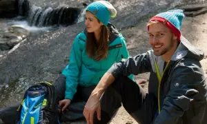 Para, co to jest trekking? Para w czapkach i kurtkach siedzi nad strumieniem, obok plecak.