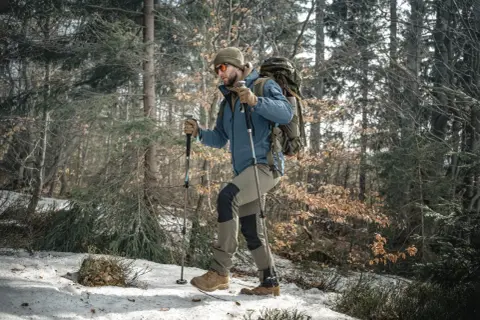 Mężczyzna w czapce i okularach przeciwsłonecznych idzie przez zaśnieżony las, wspierając się na kijach trekkingowych.