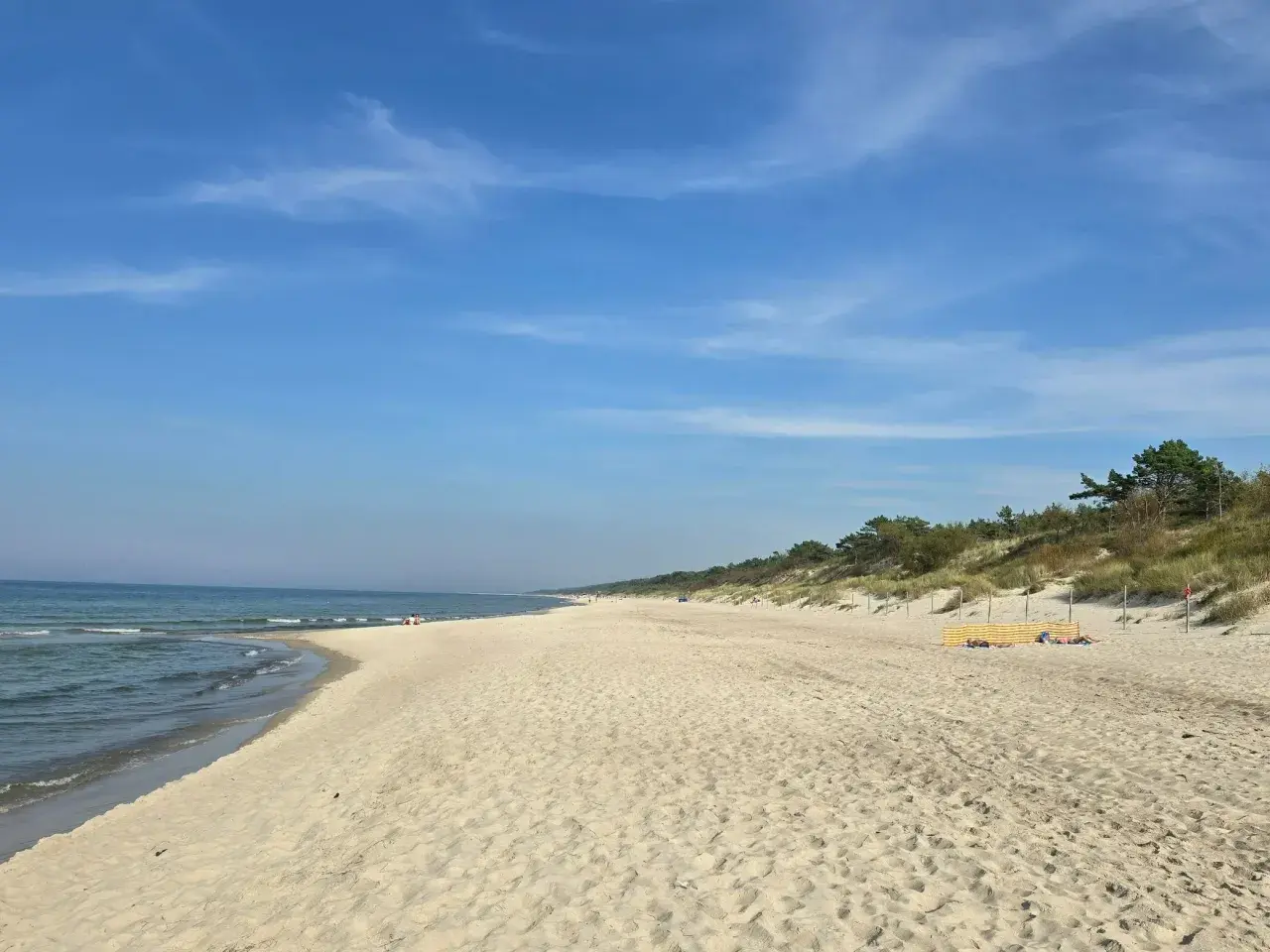 Słoneczny dzień nad zalewem kamieńskim. Szeroka, piaszczysta plaża z łagodnymi falami i zielonymi wydmami w tle. Kilka osób odpoczywa na piasku.