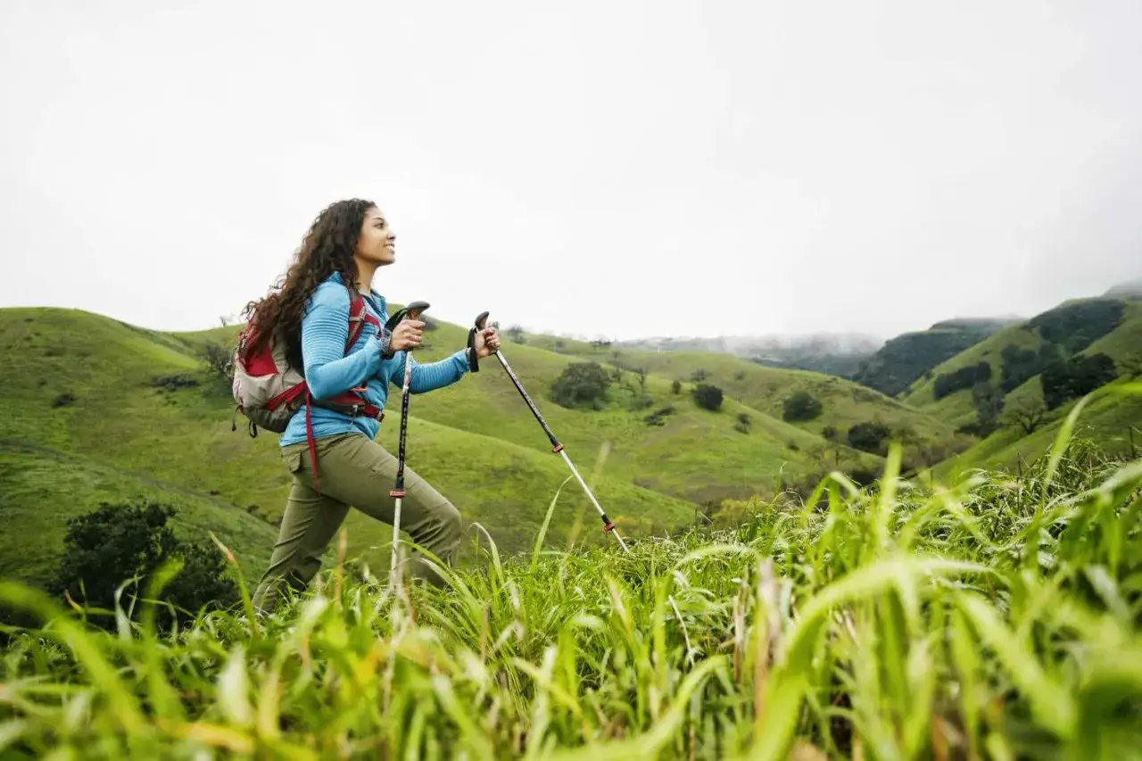 Kobieta z plecakiem i kijkami trekkingowymi, ucząca się jak chodzić z kijkami trekkingowymi, przemierza zielone wzg&oacute;rza.