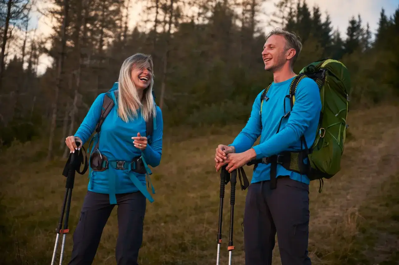 Para w turkusowej odzieży w g&oacute;ry, z plecakami i kijkami trekkingowymi, śmieje się podczas wędr&oacute;wki po lesie.
