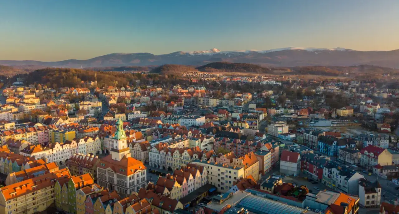 Widok na Jelenia G&oacute;rę z g&oacute;rami w tle. Kolorowe kamienice i ratusz zachęcają do odkrywania atrakcji miasta.