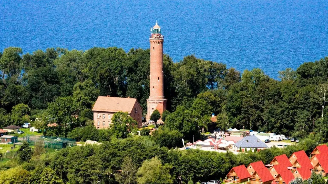 Wysoka, ceglana latarnia morska w Gąskach g&oacute;ruje nad zielonym lasem i błękitnym morzem. Obok niej budynek mieszkalny i zabudowania turystyczne.