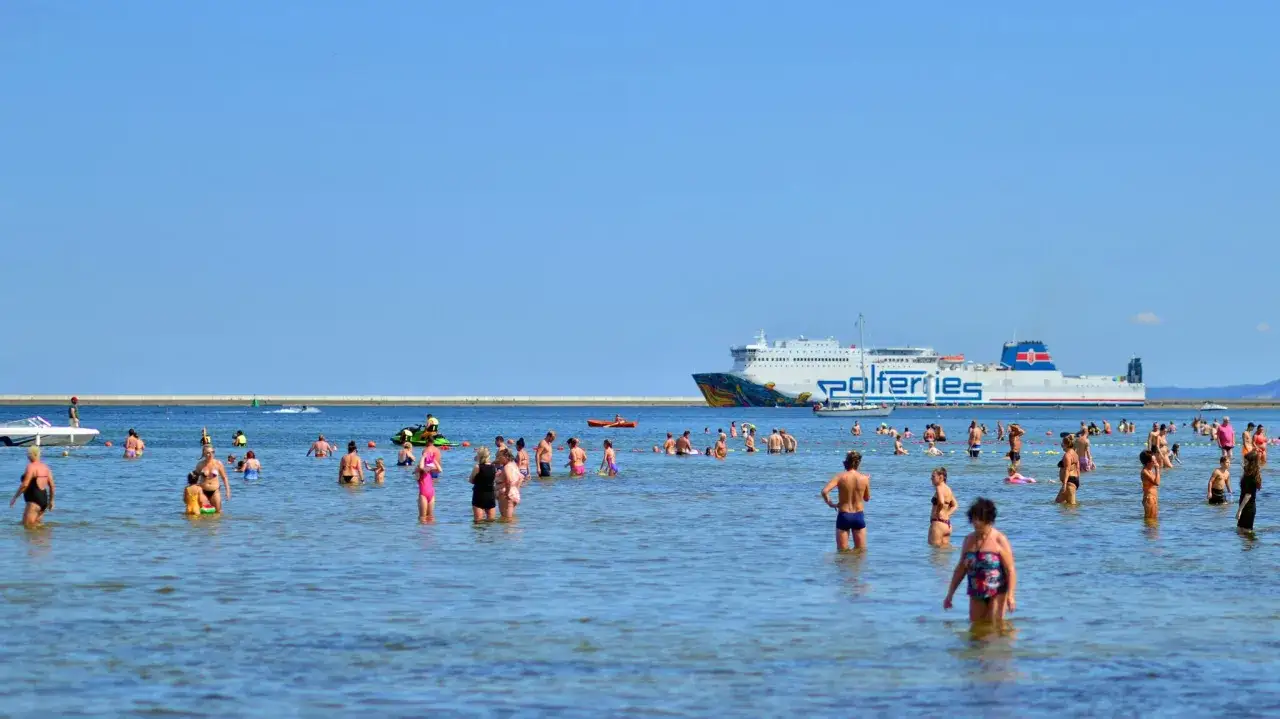 Ludzie kąpią się w morzu, a w tle płynie prom Polferries, kt&oacute;ry kursuje na trasie Świnoujście - Ystad.