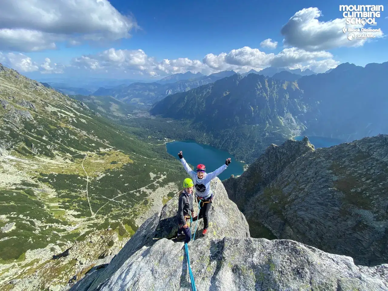 Dw&oacute;ch wspinaczy na szczycie, z widokiem na Tatry i Morskie Oko. W tle g&oacute;ry i jezioro.