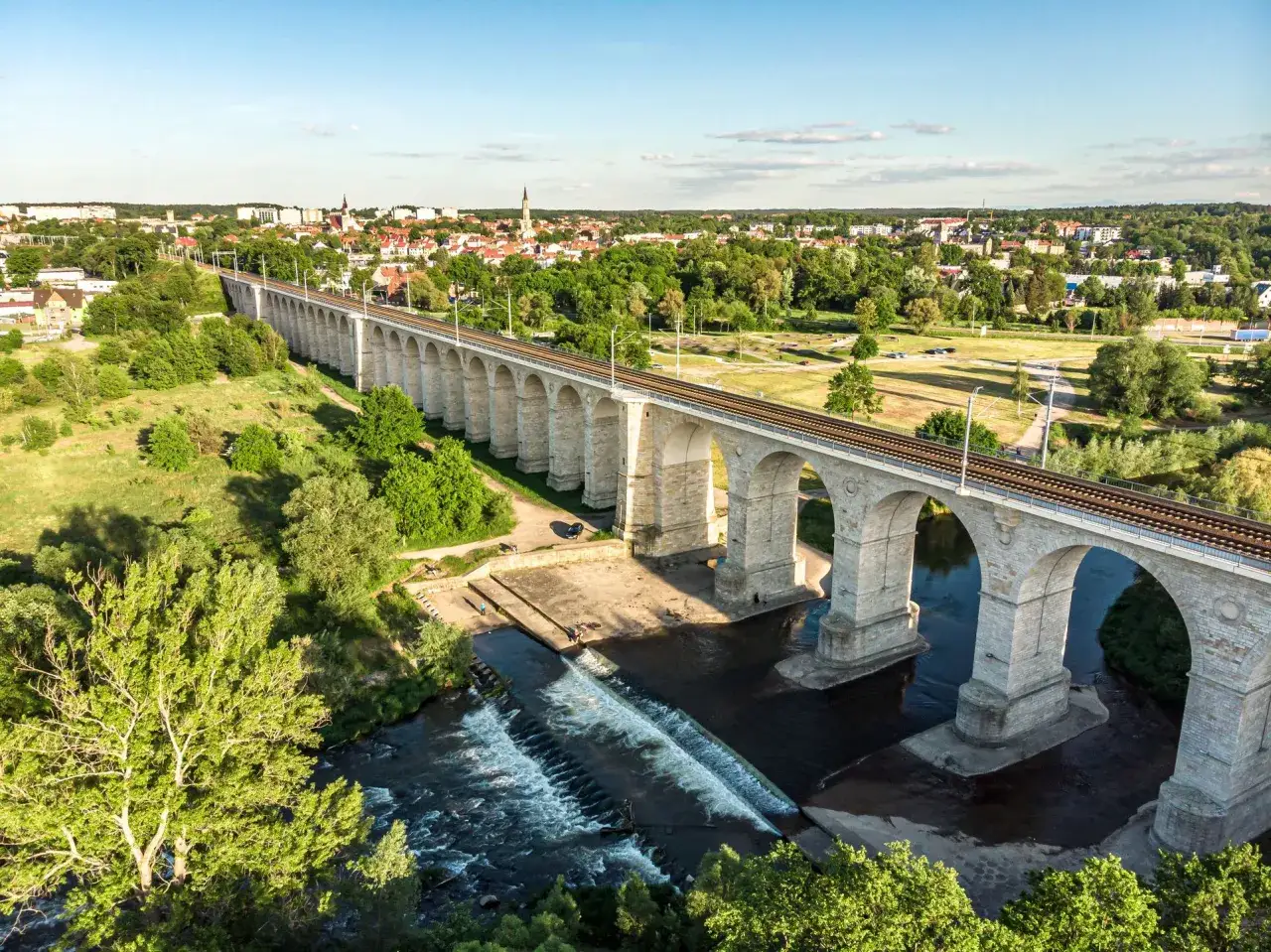 Kamienny wiadukt kolejowy nad rzeką, z miastem w tle. Jedna z gł&oacute;wnych bolesławiec atrakcje, idealna na spacer.